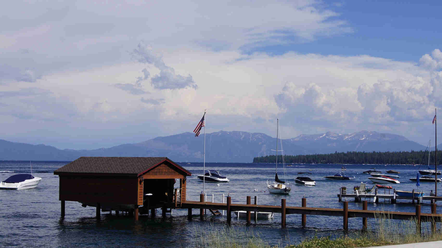 A dock with a boat house at the end, on a lake with boats, and mountains in the background