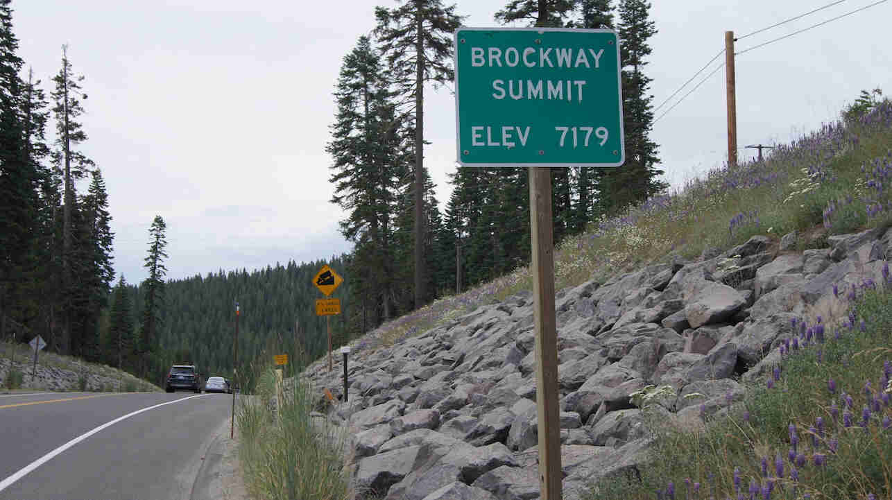 Sign showing Brockway Summit-Elev 7179, on the side of a paved road, with mountains and trees in the background