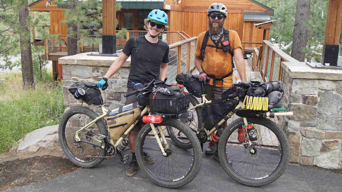 Front view of two cyclists standing with their Surly bikes, with a wood sided building in the background