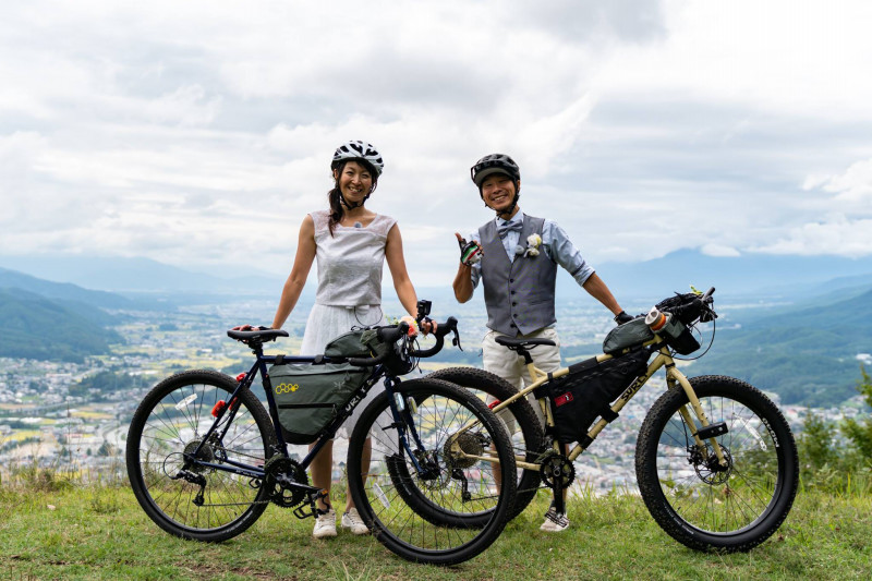Two cyclists stand with their bikes loaded with gear dressed in wedding attire at an overlook of a mountain city 