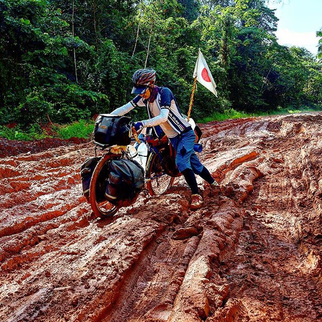 Cyclist pushing bike loaded with gear and Japan flag on back, through a deep, red mud road and forest in the background