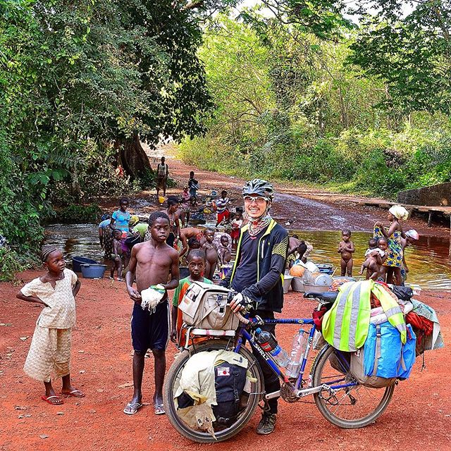 Cyclist standing on red dirt with a blue Surly bike with gear is surrounded by children in front of a forest river