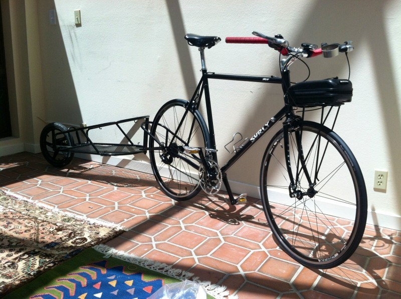 Front, right side view of a black Surly Pacer bike with trailer, parked on a terracotta floor with rugs, against a wall