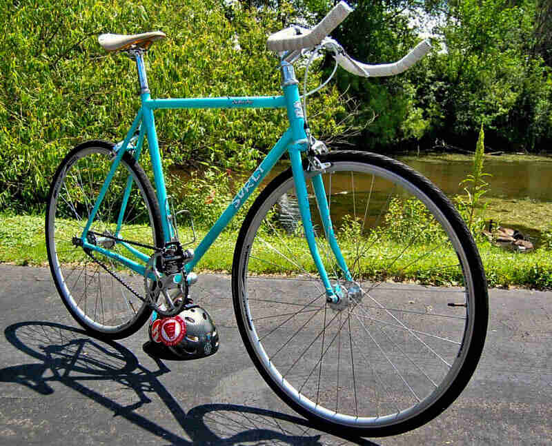 Right side view of a turquoise Surly Steamroller bike, parked on a pavement, with trees and a river in the background