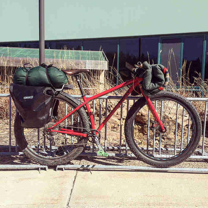 Right view of a Surly Krampus bike, red, loaded with gear, with a bike rack and building in the background