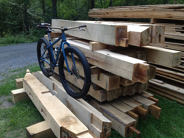 Front Right view of a Surly Krampus bike, blue, on a stack of square timbers, with a road and trees in the background