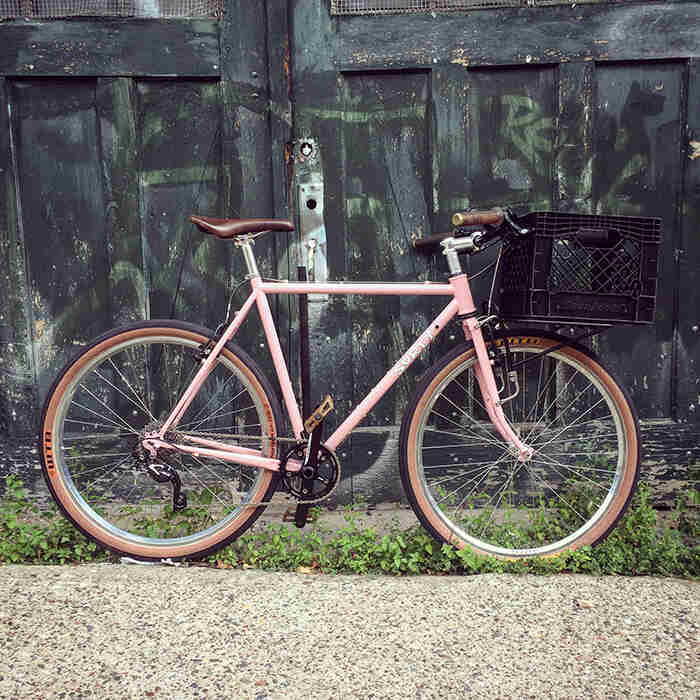 Ride side view of a pink Surly Pack Rat bike with a milk crate on front rack and old wood doors in the background