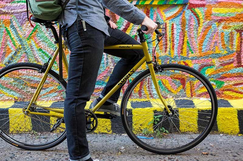 壁画の前で黄色の Surly 自転車にまたがり、片足をペダル、片足を地面において立つカジュアルな服装のサイクリスト