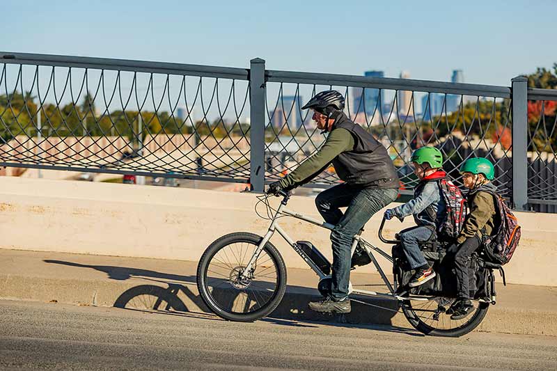 摩天楼が遠くに見えるハイウェイの橋を上を Surly Big Easy cargo 電動自転車に子供 2 人を乗せて走るサイクリスト