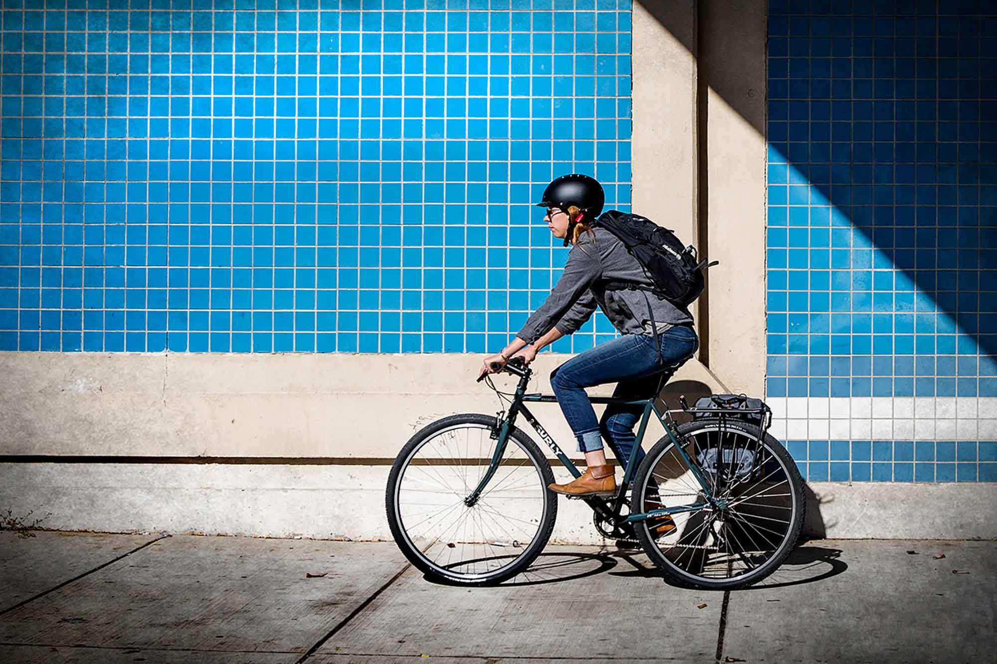 Cyclist rides a Surly bike on a sidewalk alongside a building with blue tiles on it