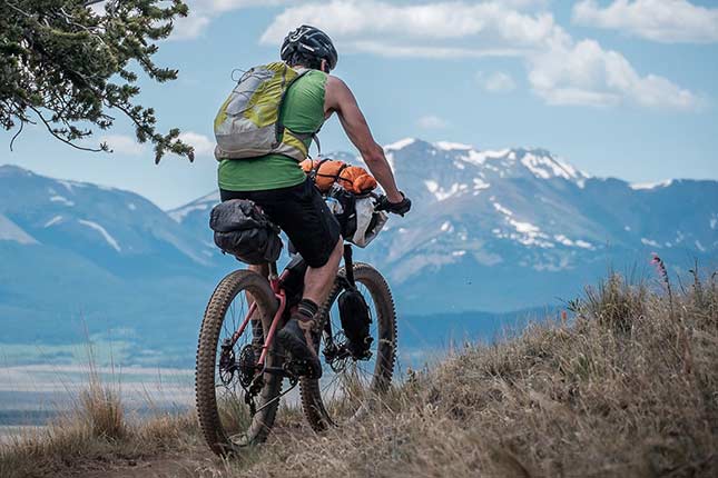 遠くに山が見える景色の中のトレイルを、ヘルメットとバックパックを身につけ、バイクパッキングギアを装着して走るマウンテンバイカー