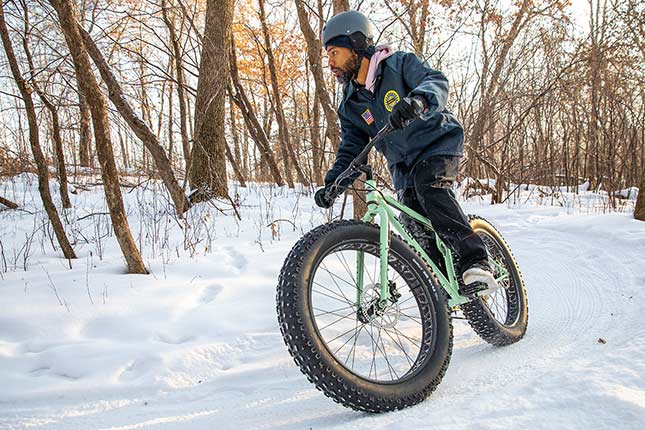 ヘルメットと防寒着を身につけてミントカラーの Surly Fat Bike に乗り雪の積もったオフロードを走るマウンテンバイカー