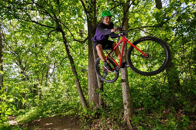 緑のヘルメットをかぶり赤い Surly Krampus に乗り、森のオフロードでジャンプするマウンテンバイカー