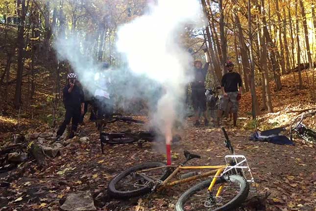 森の中の地面に倒したバイクを囲むバイカーのグループと煙を噴き出す発煙筒