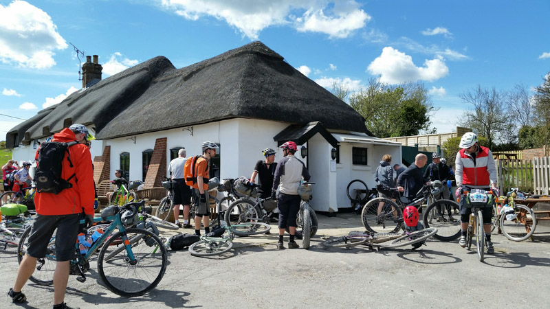 A group of cyclists, with bikes scattered around, and a building with a grass roof in the background