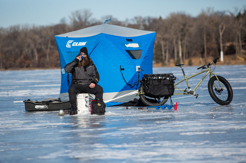 Person on a bucket on a frozen lake next to a Surly Big Fat Dummy bike, with a ice fishing shelter in the background