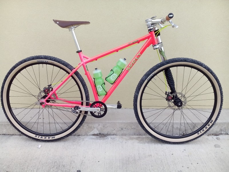 Right side view of a pink Surly Karate Monkey bike, parked next to a cement curb with a tan wall behind