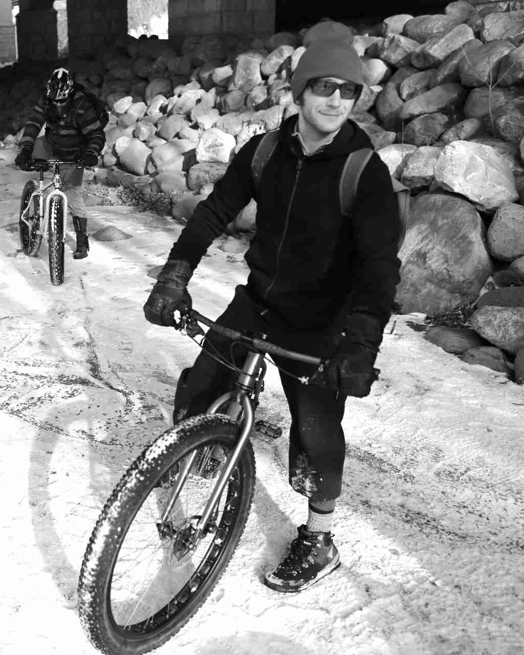 Front view of a cyclist, standing with their Surly bike on snow, with a pile of rocks behind - black & white image