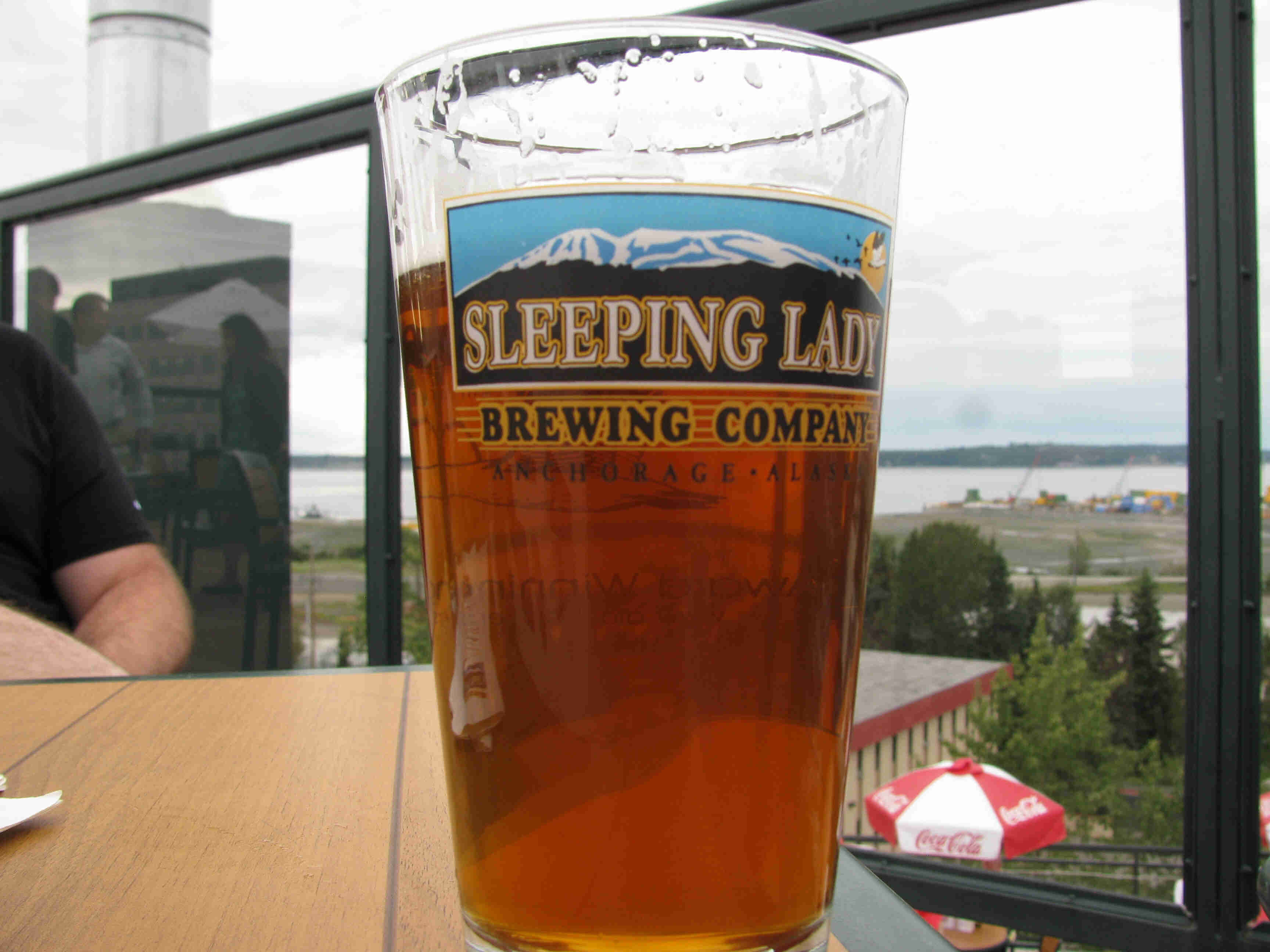 Close up view of a glass of beer, with a Sleepy Lady Brewing Company graphic, sitting on a table at an outdoor patio