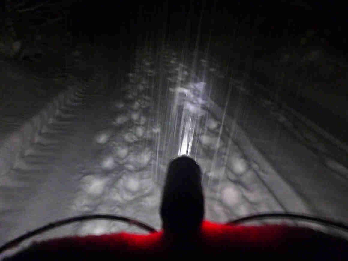 The front of the handlebar, with a headlight, on a bike that's facing down a snow covered trail at night
