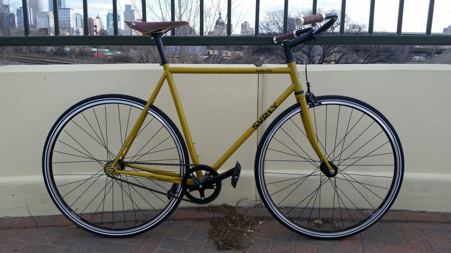 Right profile of a Surly Steamroller bike, yellow, in front of a wall on a bridge, with a city skyline in the background