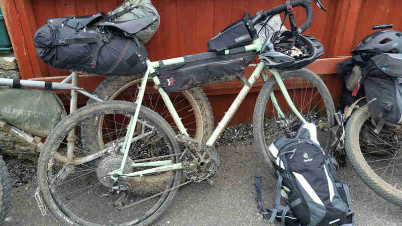 Right profile of a muddy Surly bike, loaded with gear leaning on a red wood fence