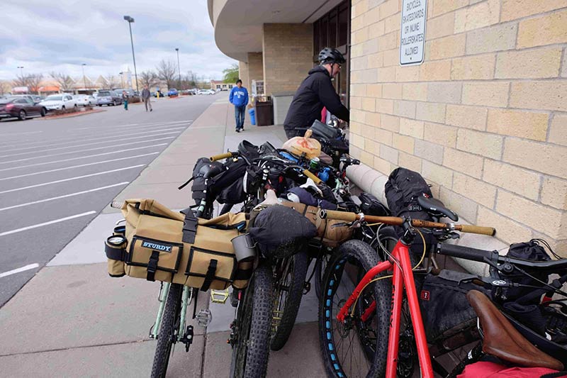 A cluster of bikes loaded with gear, next to a brick building, with a cyclist behind in the background