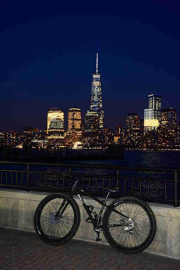 Left profile of black Surly ECR bike, parked on a sidewalk, along a rail, with a lit up NYC in the background at night