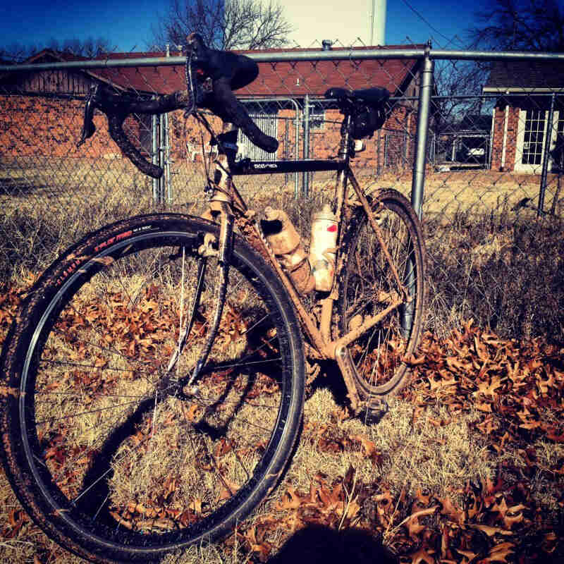 Front left view of a muddy, black Surly Cross Check bike, standing in grass, with a fenced yard of a home in background