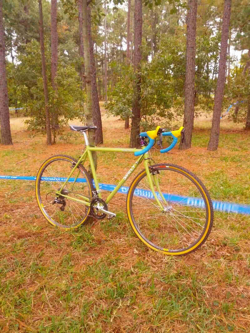 Right side view of a lime green Surly bike, leaning on a tree with race course tape wrapped around it, in a pine forest 