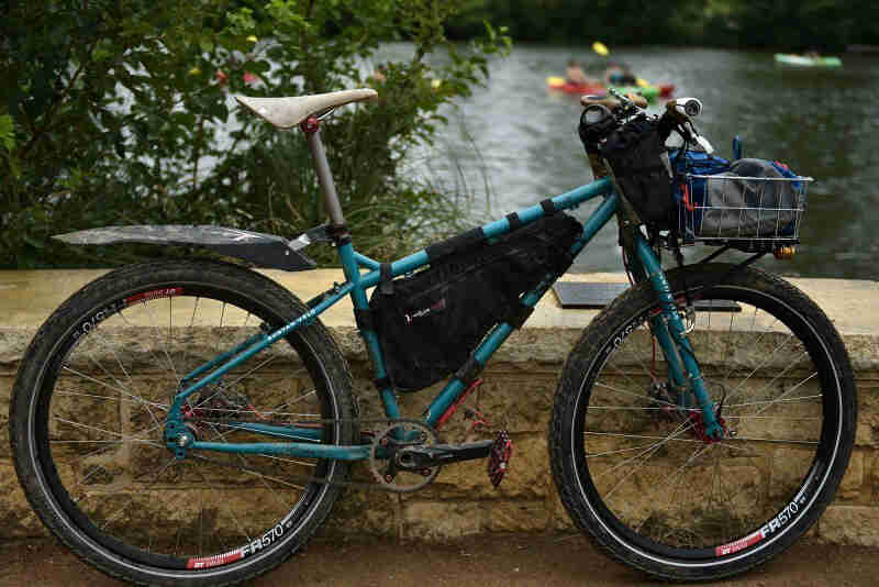 Right profile view of a turquoise, Surly Troll bike, against a stone wall, with trees and a pond in the background