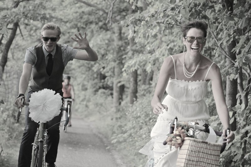 Front view of two people wearing wedding attire, riding bike down a paved trail with trees on the sides