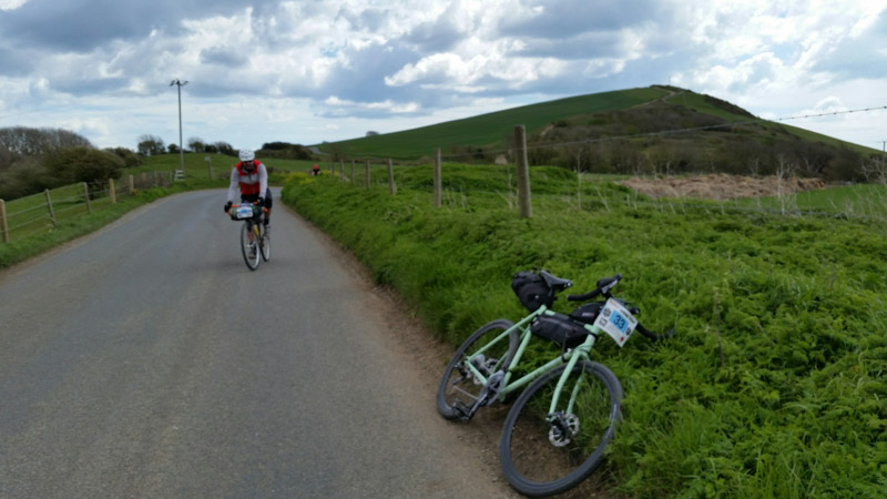 Front view of a cyclist riding down a hill on paved road with a bike on the side of the road in the grass