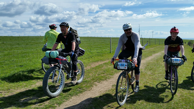 Front view of cyclists, side by side, riding down a pasture road, alongside a wire fence