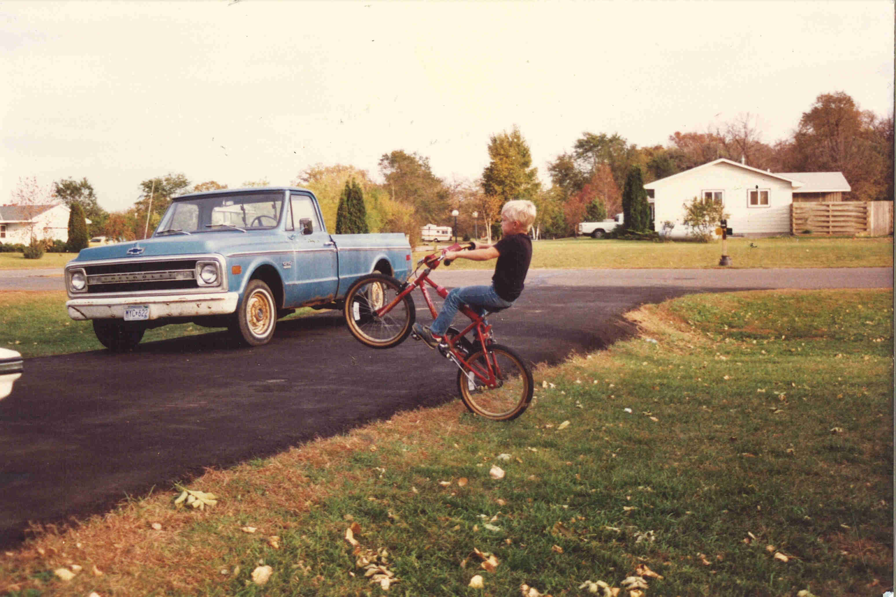 Left side view of a child riding a wheelie on a red BMX bike, next to a driveway with a blue truck parked on it