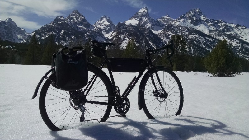 Right side view of a Surly bike with gear, parked in a field of snow, with trees and mountains in the background