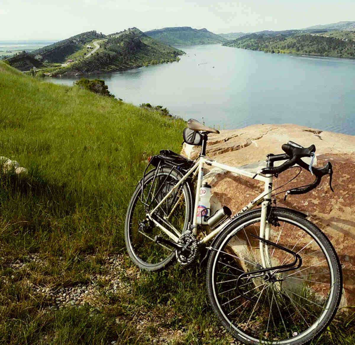 Right side view of a Surly Travelers Check bike, on a grass hilltop and leaning on a rock, with a mountain lake below