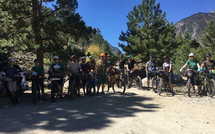 Group of cyclist lined up on a gravel patch in front of pines with mountain in the background