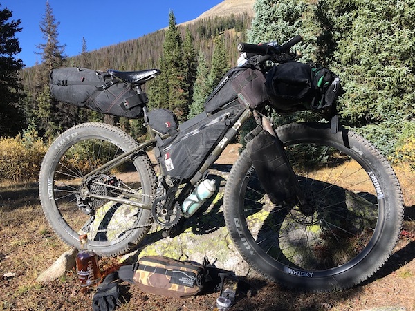 Surly bike loaded with gear leans on a rock with pine trees and a mountain hill in the background