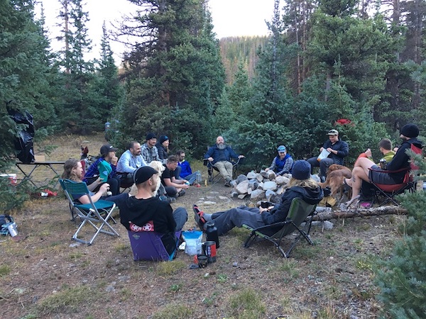 Group of people sitting in folding chairs in a circle inside of clearing surrounded by pine trees