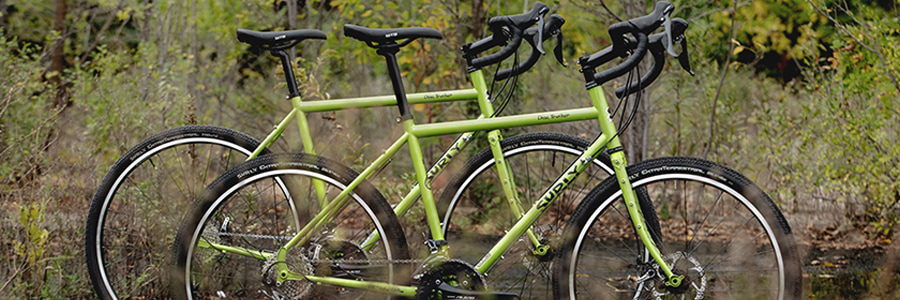 Right side view of 2, pea lime soup colored, Surly Disc Trucker bikes, side by side, on watery ground with brush behind