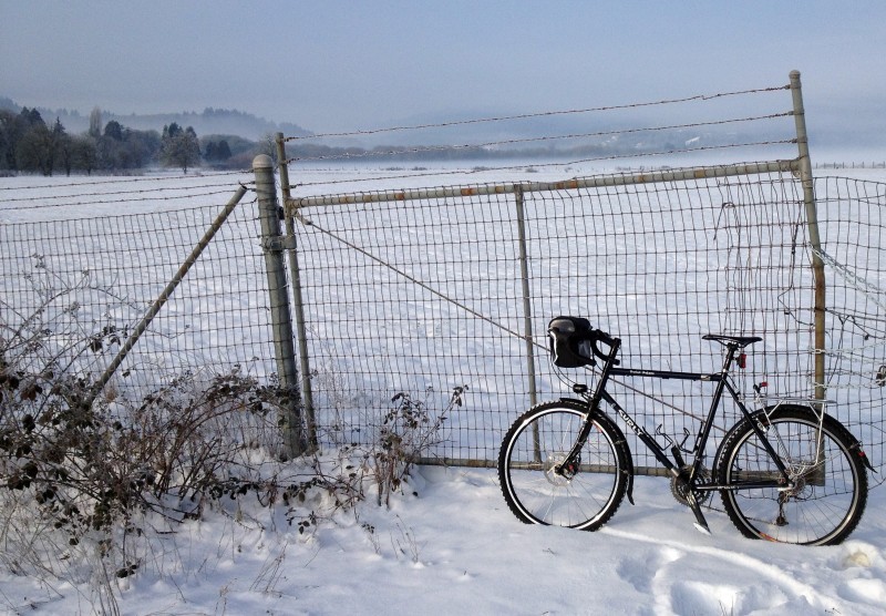 Left side view of a black Surly Long Haul Trucker bike, leaning on a wire gate, with a snow covered field behind it