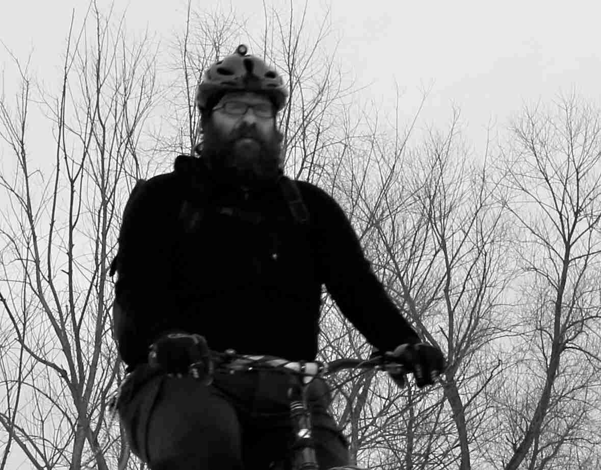 Front, waist up view of a bearded person riding a bike, with bare trees behind them - black & white image