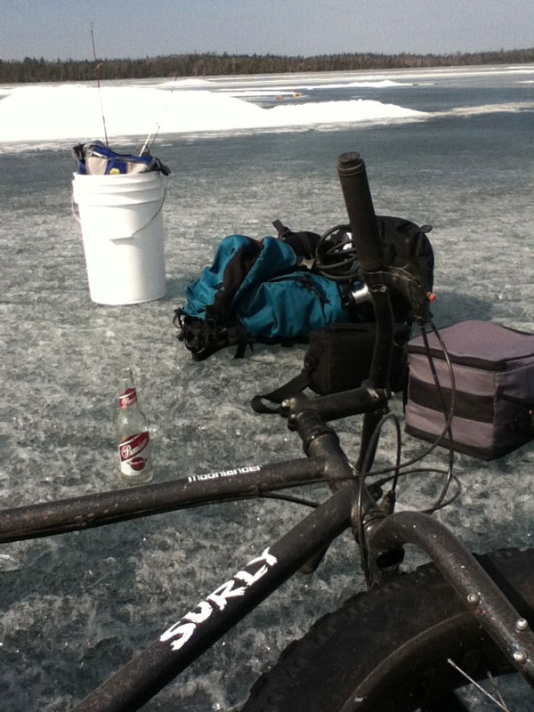 Right side view of a Surly Moonlander fat bike, laying on a frozen lake, next to ice fishing gear and a beer bottle