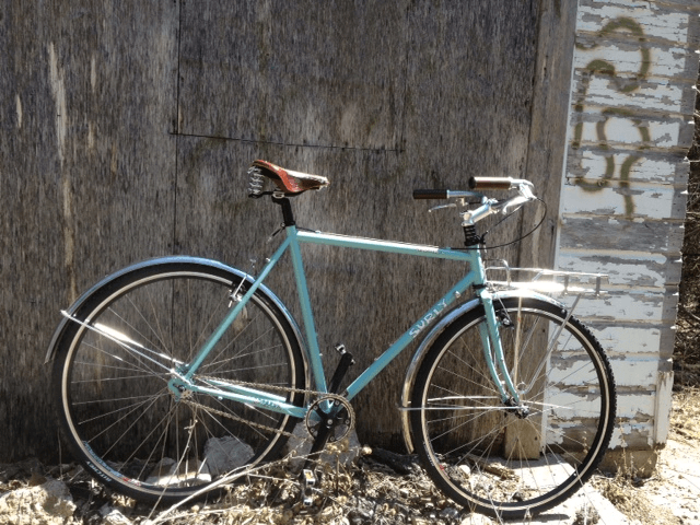 Right side view of a light blue Surly bike, leaning on an exterior, plywood wall