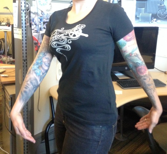 Front view of a person wearing a black Surly Steamroller, women's t-shirt, in front of a desk in an office