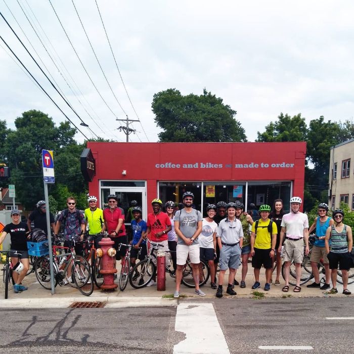 A group of cyclist stand in front red bike shop with their bicycles