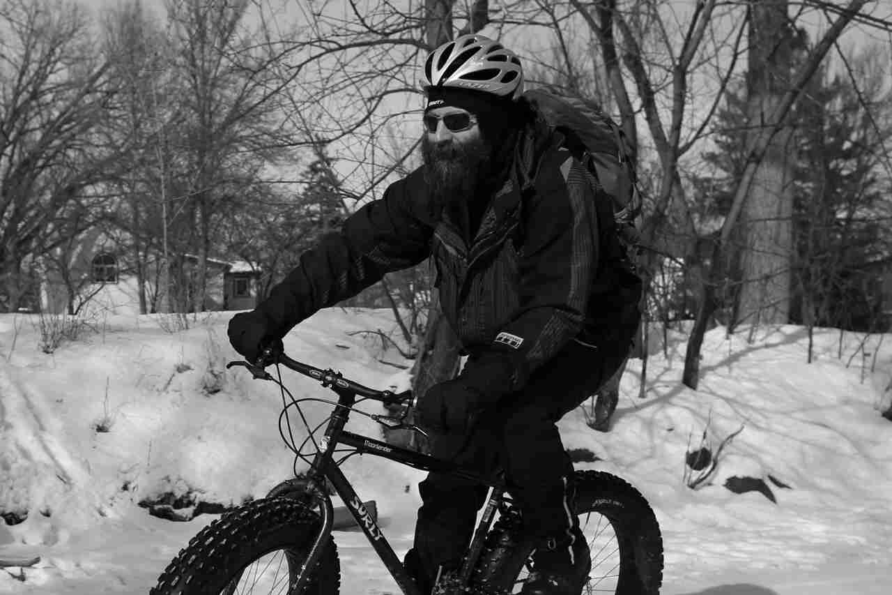 Front, left side view of a cyclist in winter attire, riding a Surly Moonlander fat bike on snow - black & white image
