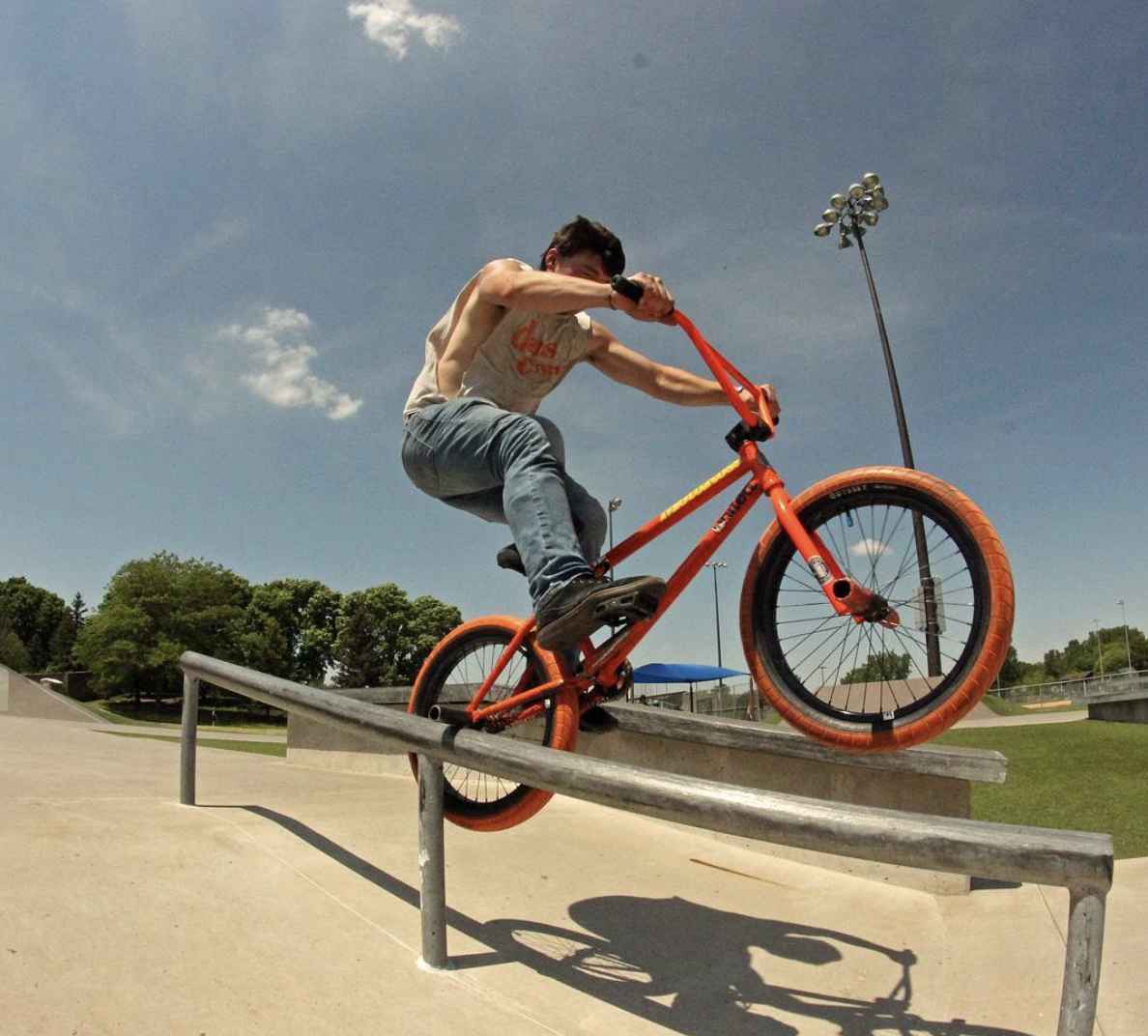 Person riding an orange BMX bike balances on the right rear axle peg down a steel rail at a rec park on a sunny day