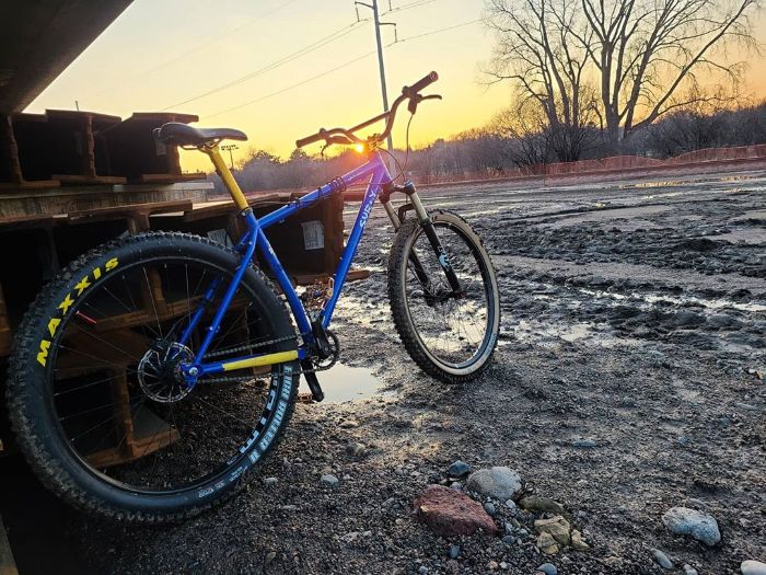 A blue colored Surly bike leans on a stack of steel beams on a muddy patch of land with the sun on the horizon